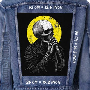 May include: A black and white illustration of a skeleton with a yellow halo praying. The image is on a denim jacket with measurements in inches and centimetres.