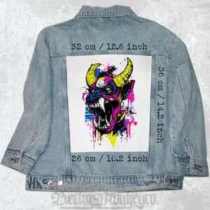 Könnte beinhalten: Eine Jeansjacke mit einem bunten Patch, der ein pinkfarbenes und gelbes Monster mit Hörnern und einem breiten Grinsen zeigt. Der Patch misst 36 cm x 26 cm.