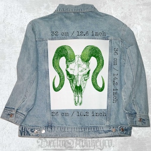 Può includere: Una toppa ricamata verde e bianca con un teschio di ariete con grandi corna. La toppa è alta 36 cm e larga 26 cm. La toppa è su una giacca di jeans blu.