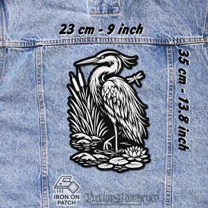 May include: Black and white iron-on patch featuring a heron standing in water with reeds, lily pads, and a dragonfly. The patch is 23 cm wide and 35 cm tall.
