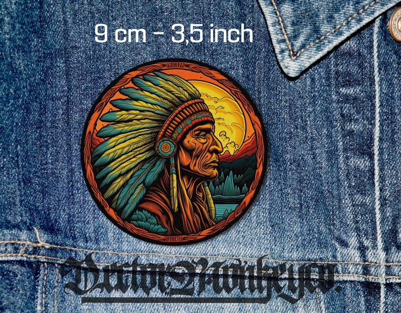 May include: A round patch featuring a Native American chief in profile, wearing a feathered headdress. The patch is 3.5 inches in diameter and set against a backdrop of a sunset and mountains. The patch is on a denim jacket.