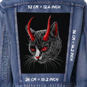 Peut inclure: Une veste en jean noire avec un patch noir représentant un chat noir et blanc avec des cornes rouges. Le patch mesure 32 cm de large, 36 cm de haut et 26 cm de profondeur. Le texte "32 CM = 12.6 INCH", "36 CM = 14.2 INCH" et "26 CM = 10.2 INCH" est imprimé sur la veste.