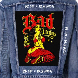 Puede incluir: Una chaqueta de mezclilla azul con un parche rectangular grande en la parte posterior. El parche muestra un gráfico de una persona con ropa roja y el texto "Bad Intentions" y "Look but don't touch". Las dimensiones son 32 cm por 36 cm por 26 cm.