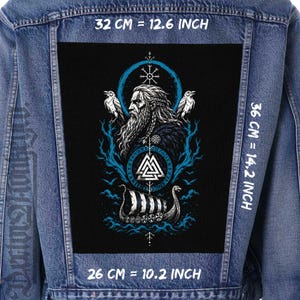 May include: A denim jacket with a large black patch depicting a Viking scene. The patch shows a bearded figure, ravens, a longboat, and the text "32 CM = 12.6 INCH", "36 CM = 14.2 INCH", and "26 CM = 10.2 INCH". The design uses blue and white.