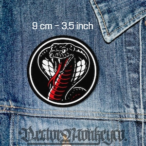 May include: A circular patch with a cobra snake design. The snake has a red tongue and white accents, set against a black background. The patch is 9 cm in diameter. The text "9 cm - 3,5 inch" is at the top, against a denim background.