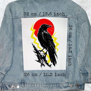May include: Light blue denim jacket with a large graphic on the back. The graphic features a black raven perched on a branch, set against a red sun and yellow lightning bolts. The graphic measures 12.6 inches wide, 14.2 inches high, and 10.2 inches long.