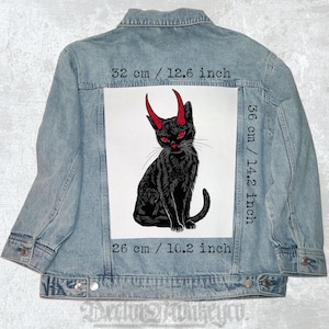 Peut inclure: Un chat noir avec des cornes rouges et des yeux rouges est assis sur un fond blanc. Le chat mesure 36 cm de haut et 26 cm de large. L'image est sur une veste en jean.