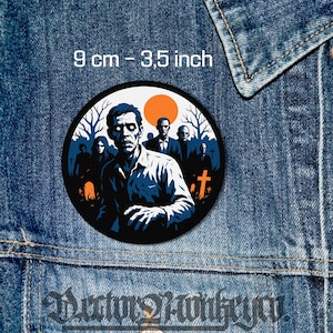 Può includere: Una toppa rotonda con un'illustrazione di zombie, 9 cm di diametro. Il disegno presenta uno zombie con una camicia blu, circondato da altri zombie, alberi e una scena di cimitero. Un sole arancione è sullo sfondo. La toppa è su una giacca di jeans.
