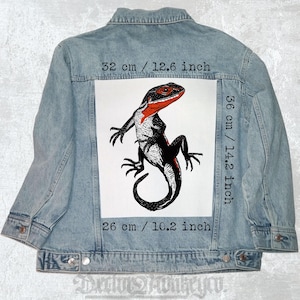 May include: Light blue denim jacket with a large white rectangular patch on the back. The patch features a black and red lizard illustration, measuring 12.6 inches wide, 14.2 inches tall, and 10.2 inches long. The jacket has a button-up front.