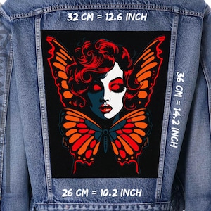 Op de afbeelding: Een zwart denim jack met een grote patch op de rug. De patch toont een vrouw met rood haar en een vlinder met oranje, rode en gele vleugels. De tekst "32 CM = 12.6 INCH", "36 CM = 14.2 INCH" en "26 CM = 10.2 INCH" is op het jack gedrukt.