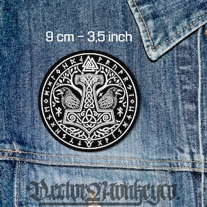 Op de afbeelding: Een ronde zwart-witte patch met een centraal ontwerp van een hamer van Thor, twee raven, een Valknut-symbool en omringende runentekst. De patch heeft een diameter van 9 cm en is afgebeeld op een achtergrond van blauwe denimstof.