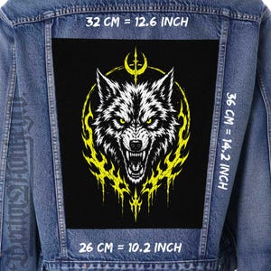 Pode incluir: Um casaco de ganga azul com um grande emblema retangular nas costas, apresentando um fundo preto, uma cabeça de lobo branca e uma borda amarela em forma de chamas. O texto indica as dimensões de 32 cm, 36 cm e 26 cm.