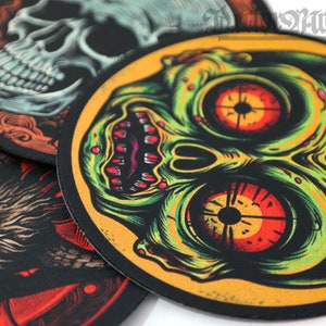 May include: A collection of round, black-edged patches with detailed illustrations. One features a skull, another a zombie-like face with green and red tones, and a third depicts a creature with red eyes. The patches have a textured appearance.