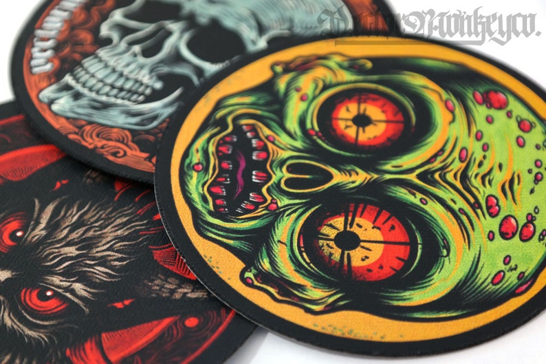 May include: A collection of round, black-edged patches with detailed illustrations. One features a skull, another a zombie-like face with green and red tones, and a third depicts a creature with red eyes. The patches have a textured appearance.