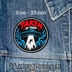 May include: Circular patch with the text "TAKEN BY THEM" in red and white. The design shows a UFO beaming a person up from a city skyline. The patch is 9 cm in diameter and is set against a denim background.
