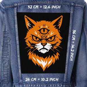 Peut inclure: Veste en jean avec un grand patch rectangulaire représentant un chat orange avec un troisième œil. Le chat a une expression fâchée et des accents blancs. Le patch mesure 32 cm de large, 36 cm de haut et 26 cm de long.