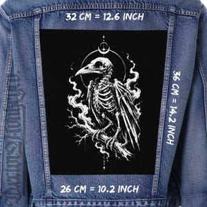 May include: A blue denim jacket featuring a large black rectangular patch on the back. The patch displays a white skeletal bird illustration perched on a branch, surrounded by smoke and a crescent moon. Dimensions are 12.6 inches by 14.2 inches by 10.2 inches.