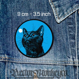 Puede incluir: Un gato negro con ojos azules mirando hacia la luna. El gato está en un círculo azul con una luna blanca en el fondo. El círculo tiene un diámetro de 9 cm.
