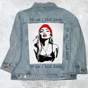 Pode incluir: Uma jaqueta de ganga com um remendo branco que apresenta uma ilustração em preto e branco de uma mulher com lábios vermelhos usando um gorro vermelho. O remendo mede 32 cm por 36 cm.