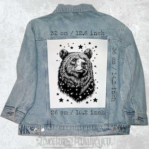 Pode incluir: Uma ilustração em preto e branco de uma cabeça de urso com estrelas ao redor. A imagem é impressa em um remendo de tecido branco, que mede 36 cm de altura e 26 cm de largura. O remendo está preso a uma jaqueta jeans azul.