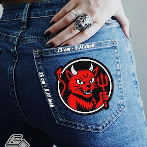 May include: A circular iron-on patch with a red devil design holding a trident, set against a black and white background. The patch is displayed on a denim pocket. The text "13 cm - 5.11 inch" is printed on the denim. The patch is labelled "IRON ON PATCH".