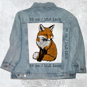 Pode incluir: Uma jaqueta de ganga com um remendo branco que apresenta uma ilustração detalhada de uma raposa vermelha sentada em um fundo branco. O remendo tem 36 cm de altura e 26 cm de largura. As medidas estão impressas na jaqueta em polegadas e centímetros.