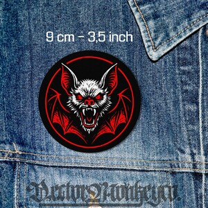 Puede incluir: Un parche redondo negro con una ilustración detallada de un murciélago con ojos y alas rojas. El murciélago es blanco con detalles rojos, sobre un fondo negro con un borde rojo. El parche mide 9 cm de diámetro.