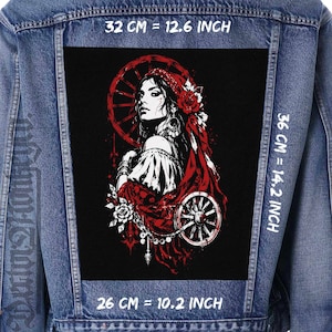 Könnte beinhalten: Eine blaue Jeansjacke mit einem großen rechteckigen schwarzen Aufnäher auf der Rückseite. Der Aufnäher zeigt eine grafische Illustration einer Person mit rotem Kopftuch, roten Rosen und Wagenrädern. Text gibt Maße von 32 cm mal 36 cm und 26 cm an.
