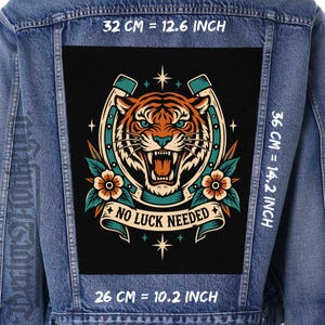 Op de afbeelding: Een blauw spijkerjack met een grote rechthoekige rugpatch met een tijgerkop, hoefijzer en bloemmotieven. De tekst NO LUCK NEEDED staat op een banner. De afmetingen zijn 32 cm breed bij 36 cm hoog, met een basis van 26 cm.