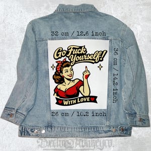 Op de afbeelding: Lichtblauwe spijkerjas met een grote afbeelding op de rug. De afbeelding toont een retro-illustratie van een vrouw in een rode jurk en rode haarband, met de tekst "Go Fuck Yourself! With Love". De afbeelding is 32 cm breed en 36 cm hoog.