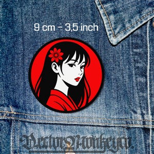 Könnte beinhalten: Ein runder Aufnäher mit einer stilisierten Illustration einer Frau mit schwarzen Haaren, roten Lippen und einer Blume im Haar, vor rotem Hintergrund. Der Aufnäher hat einen Durchmesser von 9 cm. Der Aufnäher befindet sich auf einer Jeansjacke.