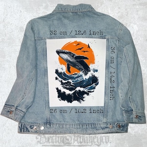 Può includere: Giacca di jeans azzurra con un disegno di balena. Il disegno mostra una balena che salta dalle onde blu, con un sole arancione e uccelli. Il disegno misura 32 cm di larghezza, 36 cm di altezza e 26 cm di lunghezza.