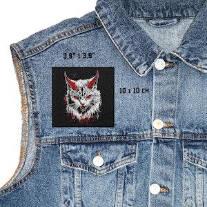 Puede incluir: Un parche cuadrado con un diseño de gato demoníaco, cuernos y ojos rojos, y detalles de sangre. El parche mide 10 x 10 cm y se muestra en una chaqueta vaquera.