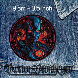 Könnte beinhalten: Ein runder, farbenfroher Patch mit einem futuristischen Roboterkopf-Design. Der Roboter hat blaue und rote Augen und ist von einem Schaltkreis-Muster umgeben. Der Patch hat einen Durchmesser von 9 cm.