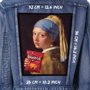Peut inclure: Veste en jean avec un grand patch représentant une parodie de "La Jeune Fille à la perle" de Vermeer. Le patch montre la jeune fille tenant un sac de Doritos. Le patch mesure 32 cm sur 36 cm et 26 cm de large.