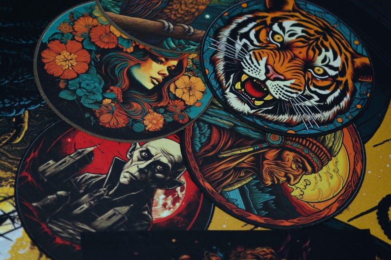 May include: Five round, colorful coasters with different designs. One features a woman with flowers, another a tiger, another a Native American, another a vampire, and the last one has a moon and stars.