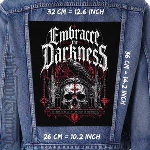 Peut inclure: Un écusson noir (26 cm x 36 cm) avec le texte "Embrace the Darkness" et un motif de crâne, de corbeau et d'architecture gothique. L'écusson est sur une veste en jean. Le texte "Nothing is real - Everything is permitted" est également présent.