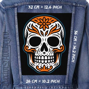 Op de afbeelding: Spijkerjasje met een groot, zwart embleem met een suikerschedelontwerp voor de Dag van de Doden. De schedel is wit met oranje en teal accenten en meet 32 cm breed en 36 cm hoog.
