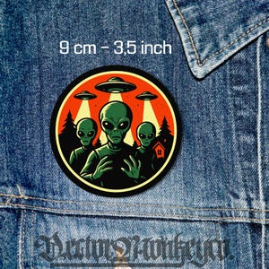 Op de afbeelding: Een ronde patch met drie groene aliens en vliegende schotels. De patch, met een diameter van 9 cm, heeft een oranje en zwarte rand met een silhouet van een bos en een huis. De tekst op de patch luidt "9 cm - 3,5 inch".