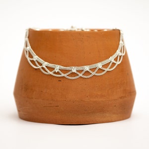 Puede incluir: Un tobillo de plata con un diseño delicado de cadena y pequeños acentos redondos. El tobillo se muestra en una maceta de terracota.