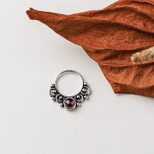 May include: Silver nose ring with a decorative design and a small, round, dark red gemstone in the center.