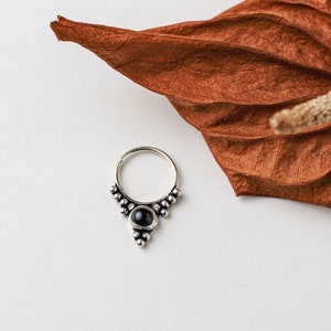 May include: A silver septum ring with a black gemstone center and decorative beaded accents. The jewelry is set against a white background with a dried, brown leaf in the upper right corner. The ring is a piece of body jewelry.