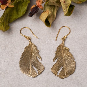 Puede incluir: Un par de pendientes dorados con forma de pluma. Cada pendiente presenta un diseño detallado de pluma con un gancho. Los pendientes se muestran sobre una superficie gris claro, con follaje seco en el fondo.
