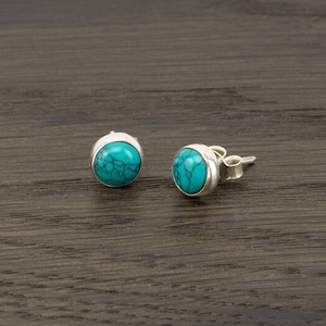 Puede incluir: Un par de pendientes de botón con piedras redondas de color turquesa engastadas en plata. Las piedras turquesas tienen un patrón veteado natural. Los pendientes se exhiben sobre una superficie de madera oscura.