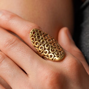 Könnte beinhalten: Ein goldener Ring mit einem detaillierten, komplizierten Design aus wirbelnden Mustern. Der Ring ist oval geformt und hat ein filigranes, durchbrochenes Design.