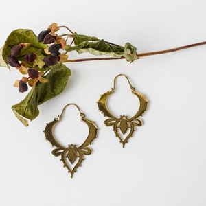 Puede incluir: Un par de pendientes de color latón con un diseño calado ornamentado. Los pendientes tienen un motivo floral o de hoja y se exhiben sobre una superficie blanca con follaje seco.
