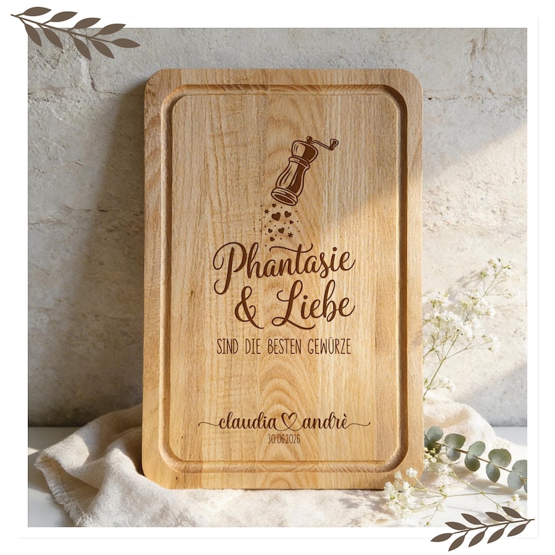 Op de afbeelding: Een rechthoekige houten snijplank met een gegraveerde rand. Het bord heeft gegraveerde tekst en een pepermolenillustratie. De tekst luidt "Phantasie & Liebe sind die besten Gew&uuml;rze" en bevat namen en een datum.