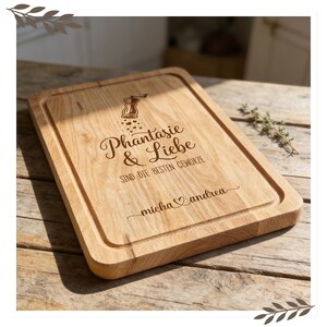 Op de afbeelding: Een rechthoekige houten snijplank met een verzonken rand. De plank heeft gegraveerde tekst en een afbeelding van een kruidenmolen. De tekst luidt "Phantasie & Liebe sind die besten Gew&uuml;rze" en bevat de namen "Micha" en "Andrea".
