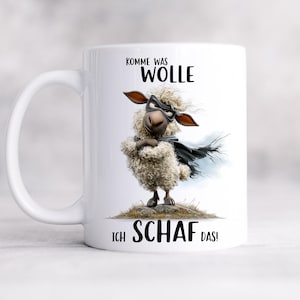 Könnte beinhalten: Weiße Keramiktasse mit einem Comic-Schaf als Superheld, mit Maske und Umhang. Auf der Tasse ist der deutsche Text "KOMME WAS WOLLE ICH SCHAF DAS!" aufgedruckt.