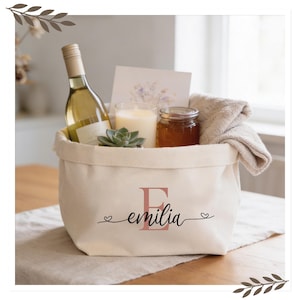 Könnte beinhalten: Ein cremefarbener Geschenk-Korb aus Canvas mit dem Namen "emilia" auf der Vorderseite. Der Korb enthält eine Flasche Weißwein, eine Kerze, ein Glas Honig, eine Sukkulente, eine Karte und ein gefaltetes Handtuch. Der Buchstabe "E" ist rosa gedruckt.